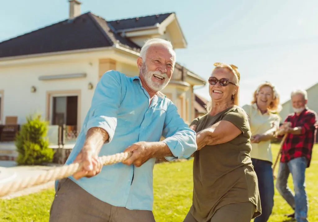 Two senior adults joyfully playing tug of war with a rope in a sunny backyard, with another couple in the background.