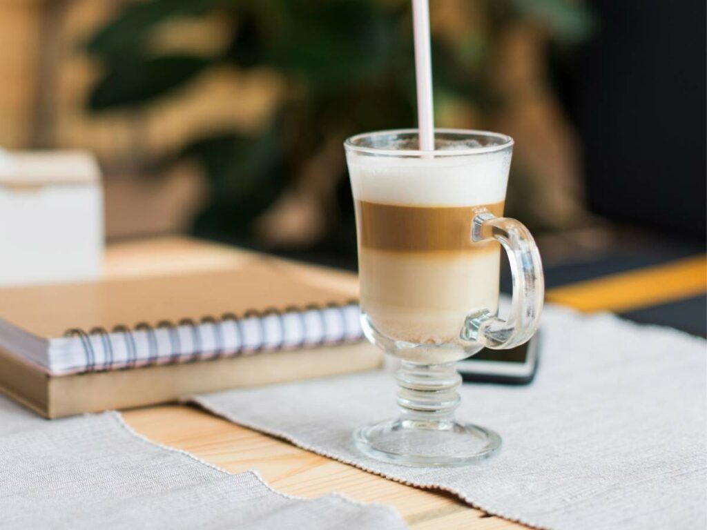A layered latte in a clear glass with a handle sits on a table next to a spiral notebook and other items.