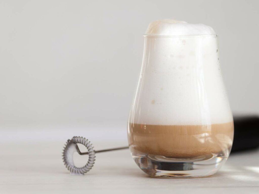 A glass cup filled with a layered drink topped with frothy foam, sitting next to a small, handheld whisk on a light-colored surface.