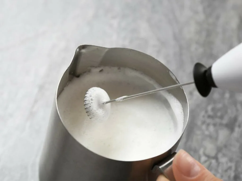 A hand froths milk in a metal pitcher using a handheld electric frother.