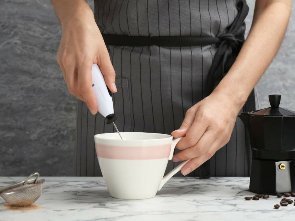 Person using a handheld milk frother in a white mug on a marble countertop. A coffee pot and a sieve with coffee beans nearby.
