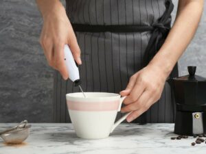 Person using a handheld milk frother in a white mug on a marble countertop.