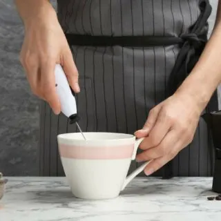 Person using a handheld milk frother in a white mug on a marble countertop.