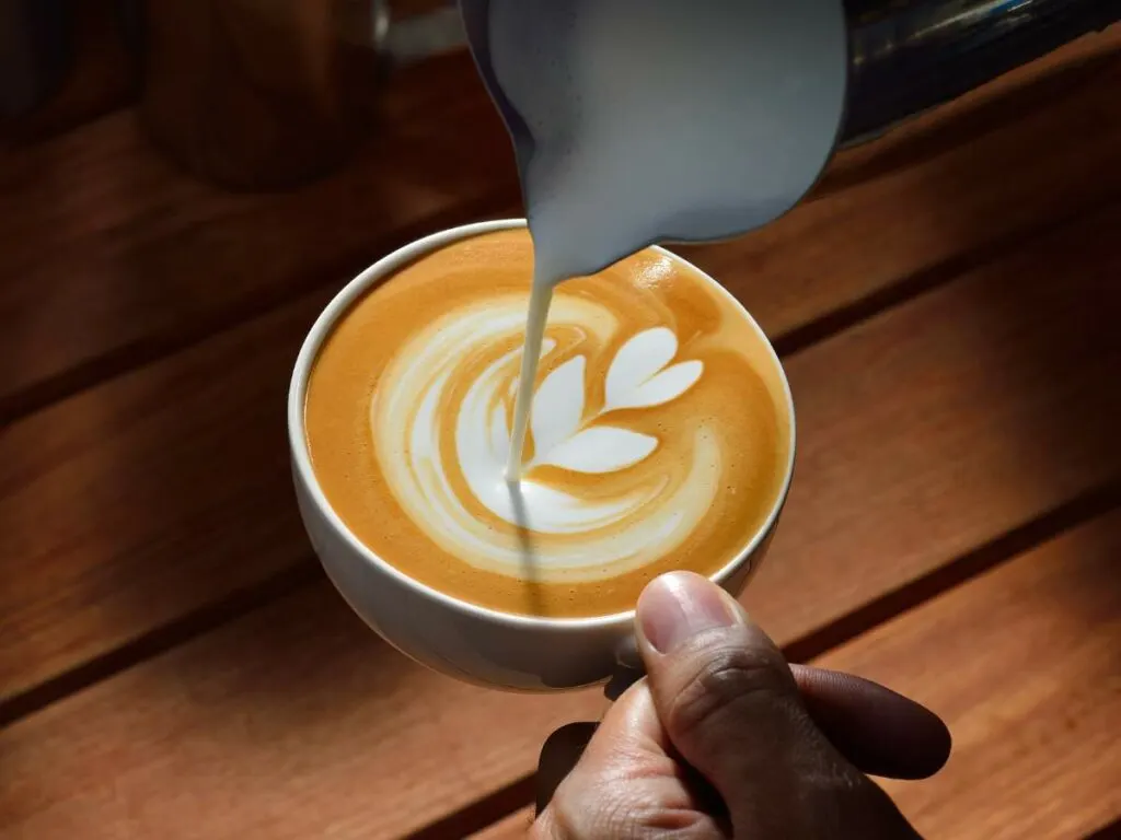 A hand holding a cup of latte with foam art while milk is being poured into it. The cup is on a wooden table.
