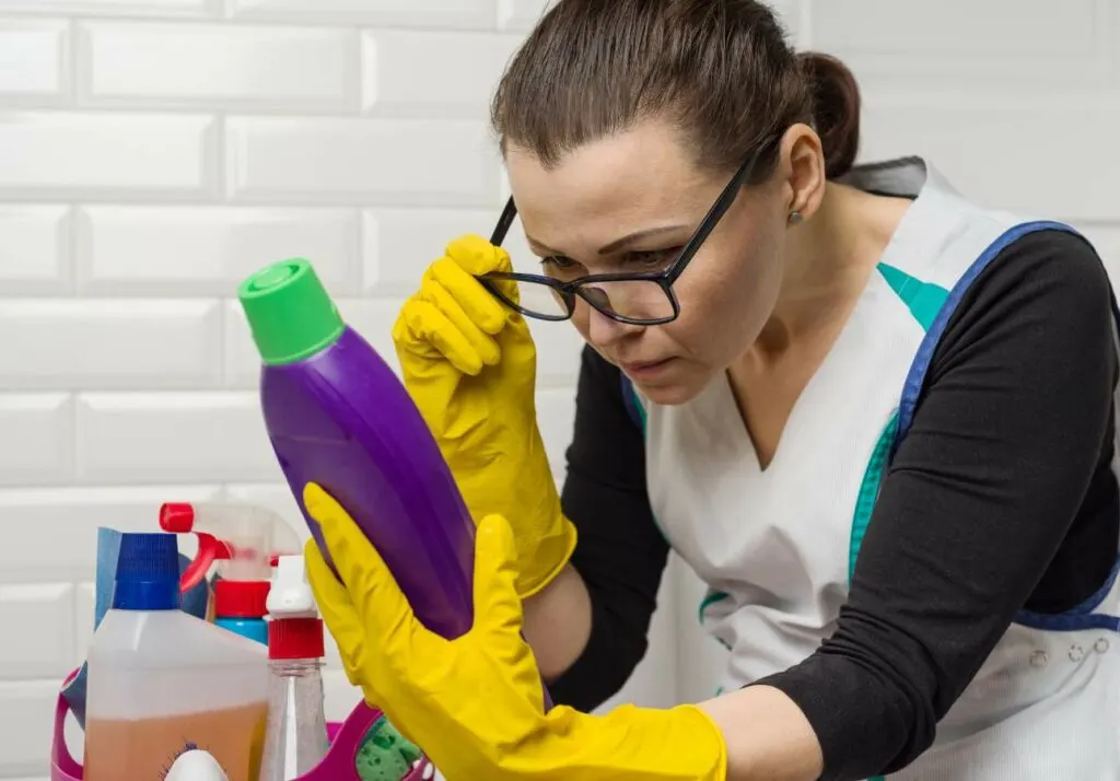 A woman wearing yellow gloves and glasses closely examines a bottle of cleaning product. Several other cleaning supplies are visible in the foreground.