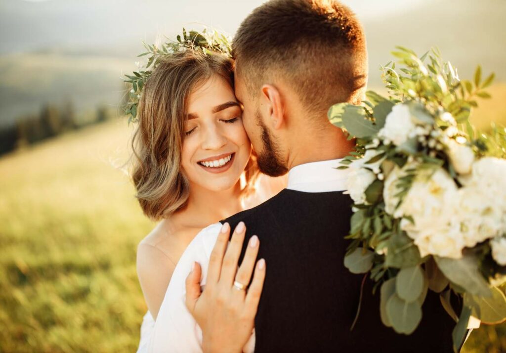 A bride and groom embrace in a grassy field, the bride smiling with closed eyes while holding a bouquet, and the groom facing away from the camera.