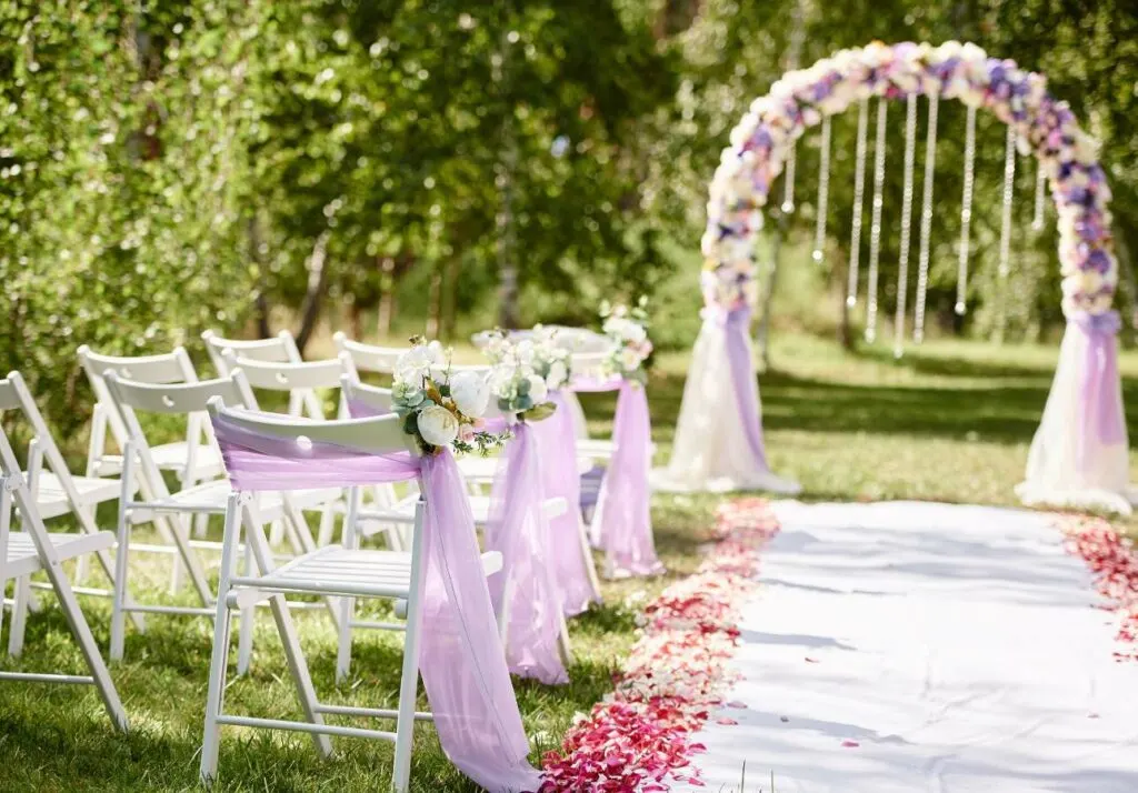 Outdoor wedding ceremony setup with white folding chairs draped in lavender fabric, a flower-adorned arch, and a petal-strewn white aisle runner in a garden setting.