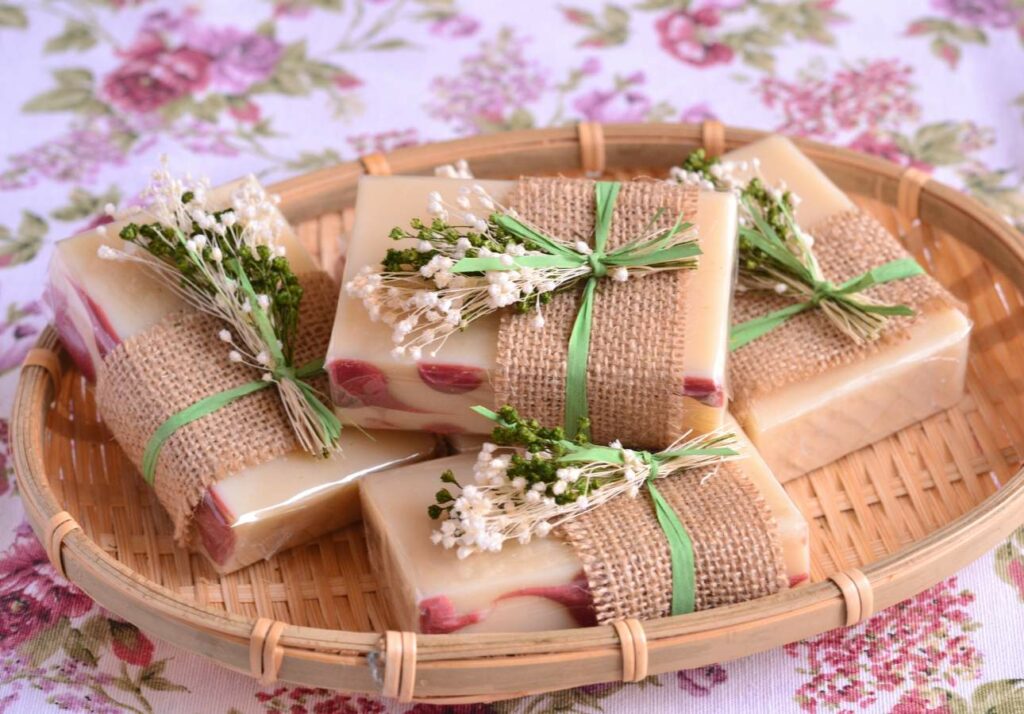 Handmade soap bars wrapped in burlap and decorated with small sprigs of dried flowers arranged in a wicker basket on a floral tablecloth.