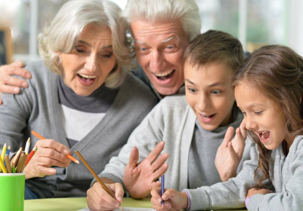 Two elderly adults and two young children excitedly engage in a drawing activity at a table.