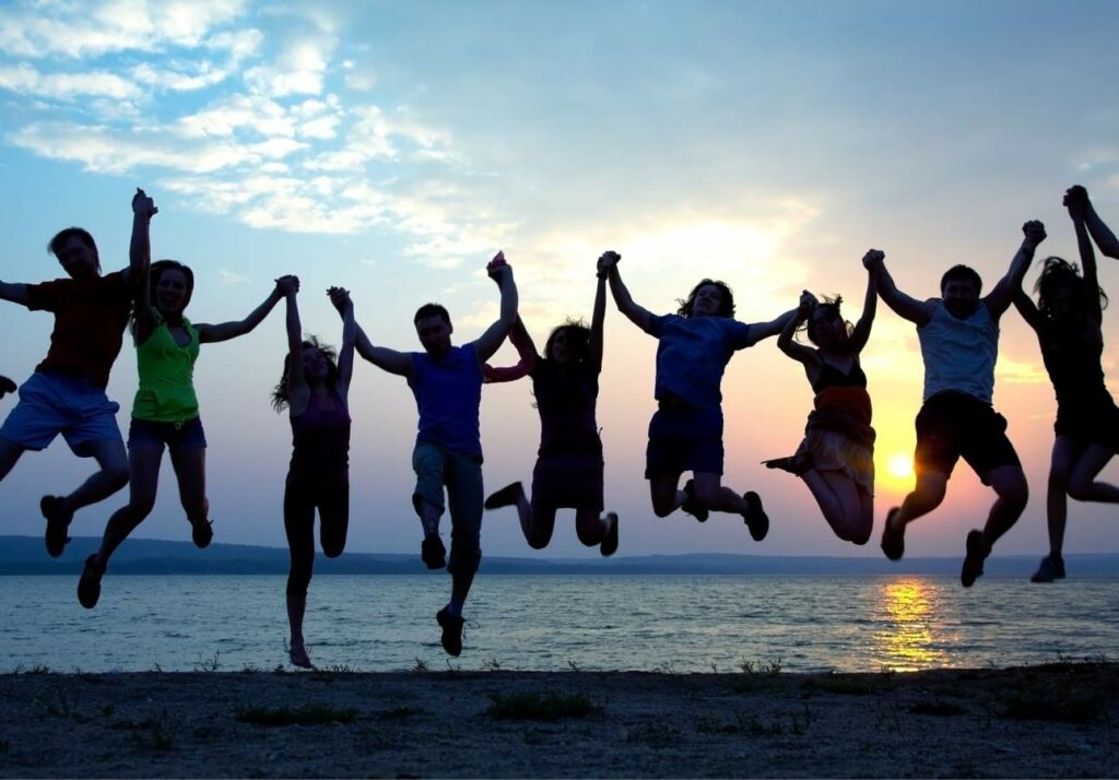 A group of eight people joyfully jumping in silhouette against a sunset over a large body of water.