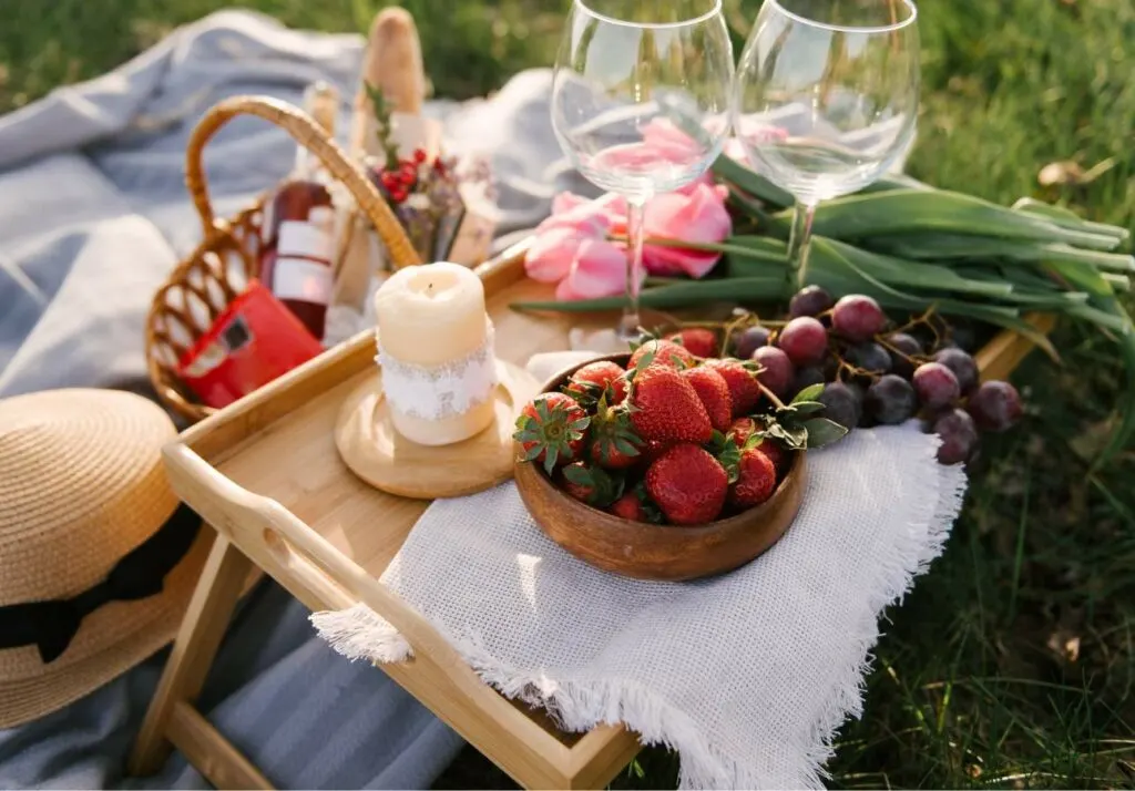 A romantic picnic setup on the grass featuring a basket, wine glasses, strawberries, grapes, bread, and a lit candle on a small tray.
