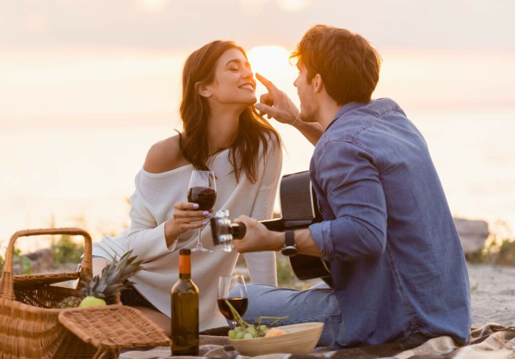 A couple enjoys a romantic sunset picnic on the beach, with a man tenderly touching a woman's face as they sip wine.