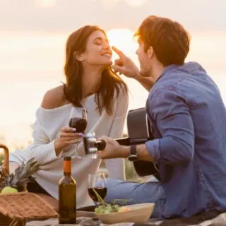 A couple enjoys a romantic sunset picnic on the beach, with a man tenderly touching a woman's face as they sip wine.