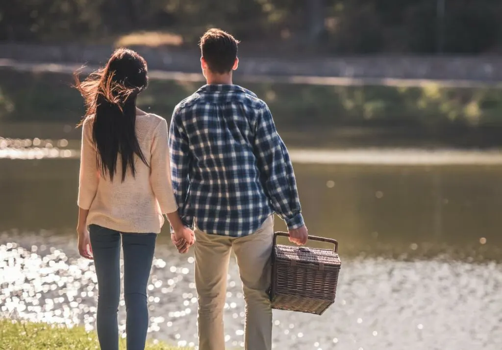 A couple holding hands and carrying a picnic basket by a lake, dressed in casual attire, walking away from the camera.