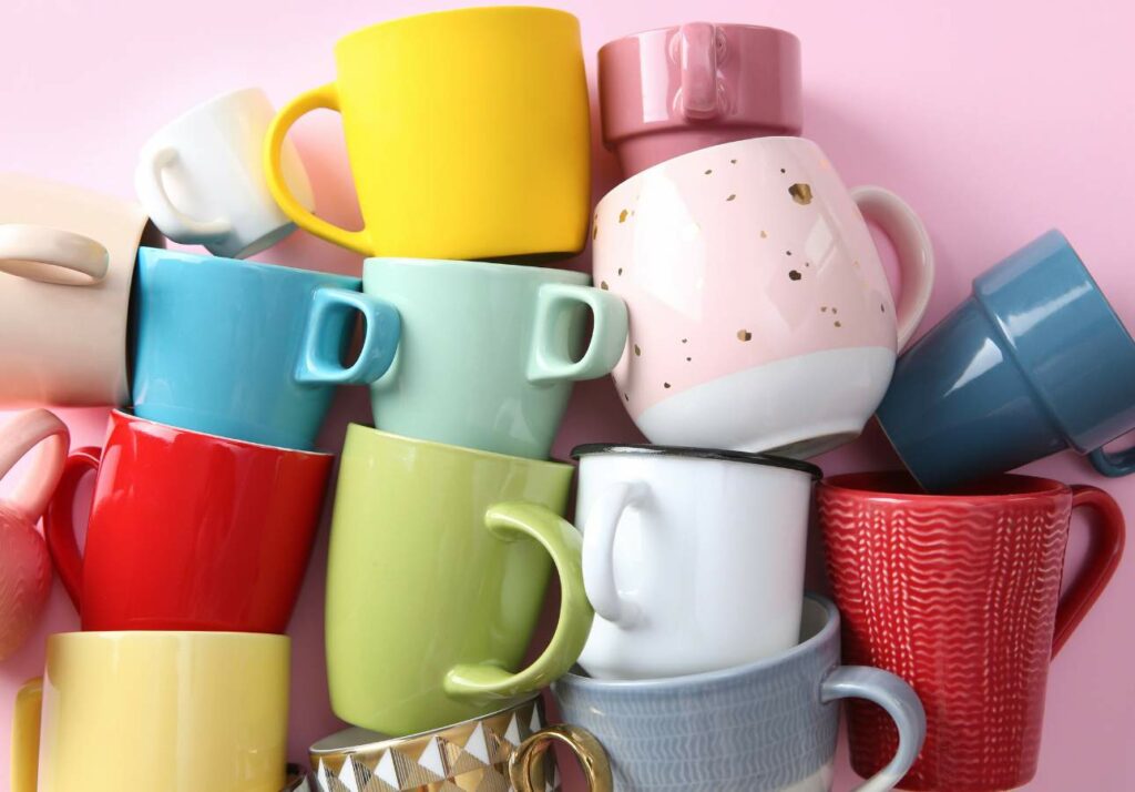 A variety of colorful mugs in different sizes and designs piled together on a pink background.