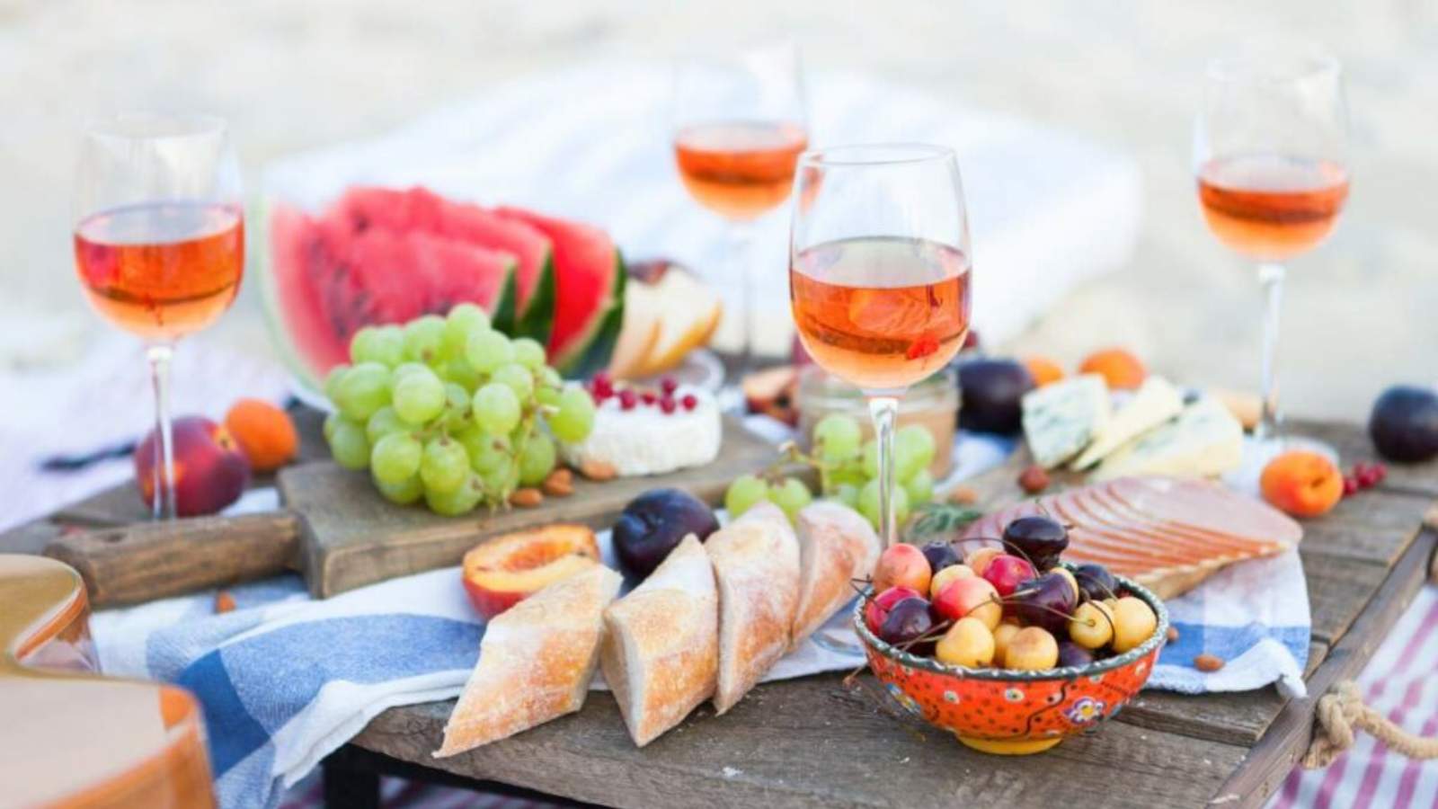 A beach charcuterie board with glasses of rosé wine, a variety of fruits and sliced baguette.