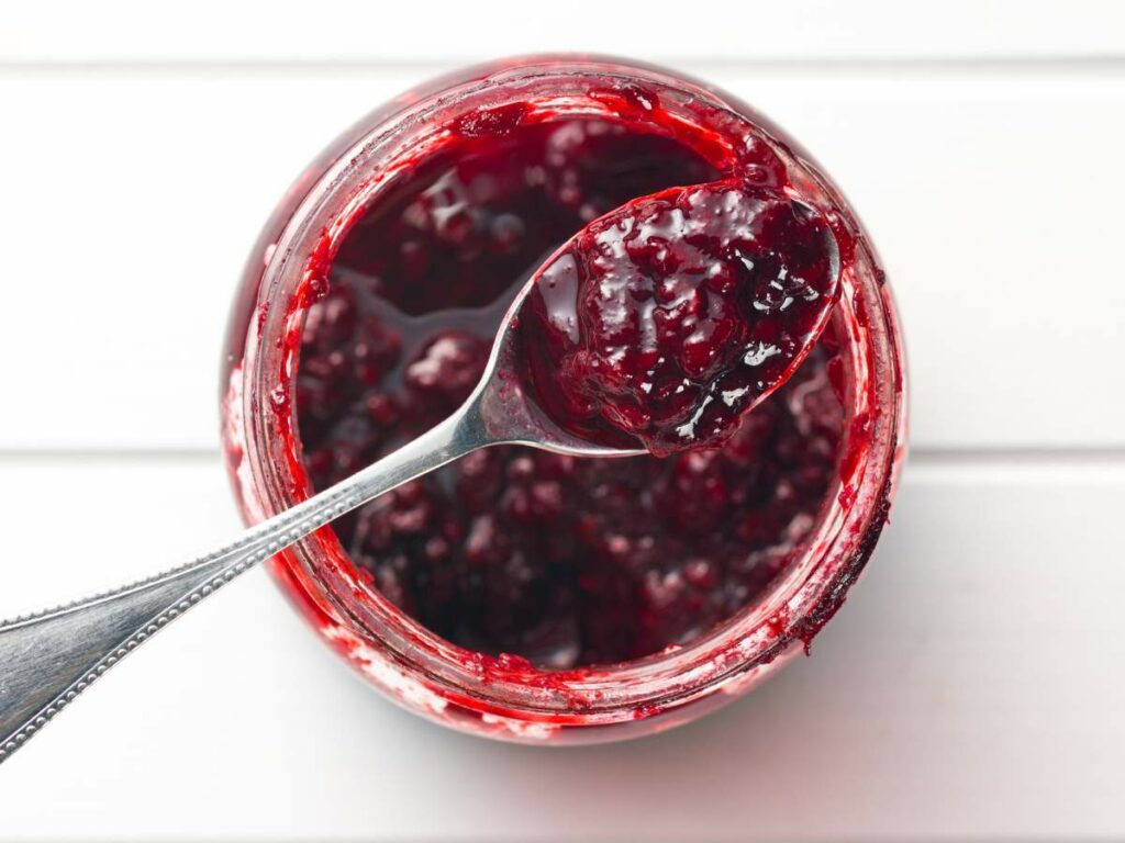 A spoon rests inside a jar filled with dark red raspberry jam, with some jam on the spoon.