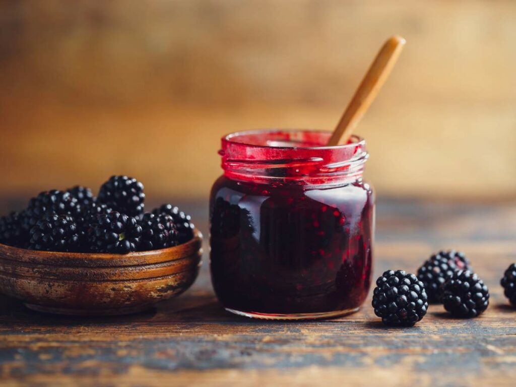 A glass jar filled with blackberry jam with a wooden spoon inside it.