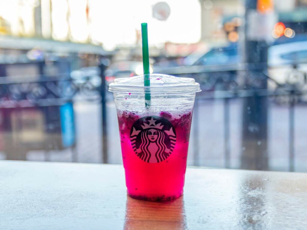 A clear plastic cup with a green straw, containing a pink-colored drink with ice, sits on a wooden table.