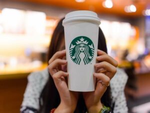 Person holding a white Starbucks coffee cup with the Starbucks logo, in front of their face.