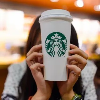 Person holding a white Starbucks coffee cup with the Starbucks logo, in front of their face.