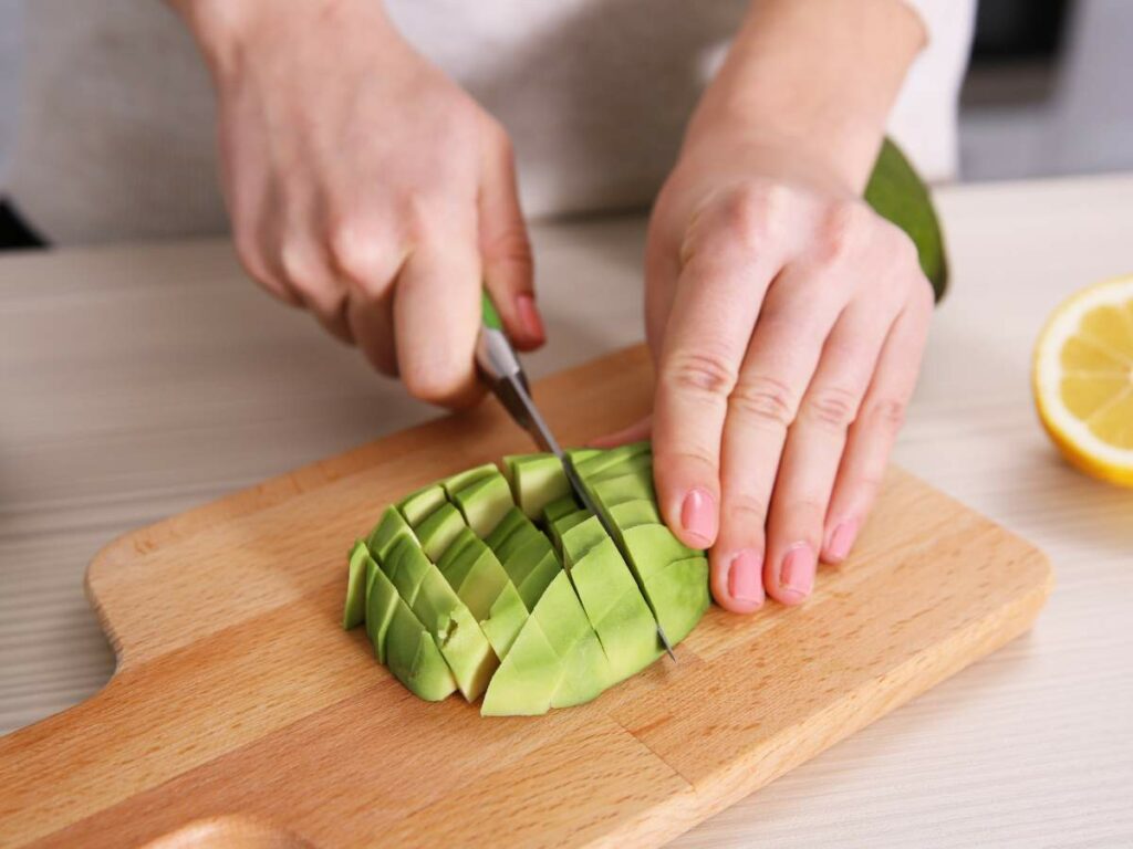 A person is slicing an avocado on a wooden cutting board. A lemon is also visible on the side.