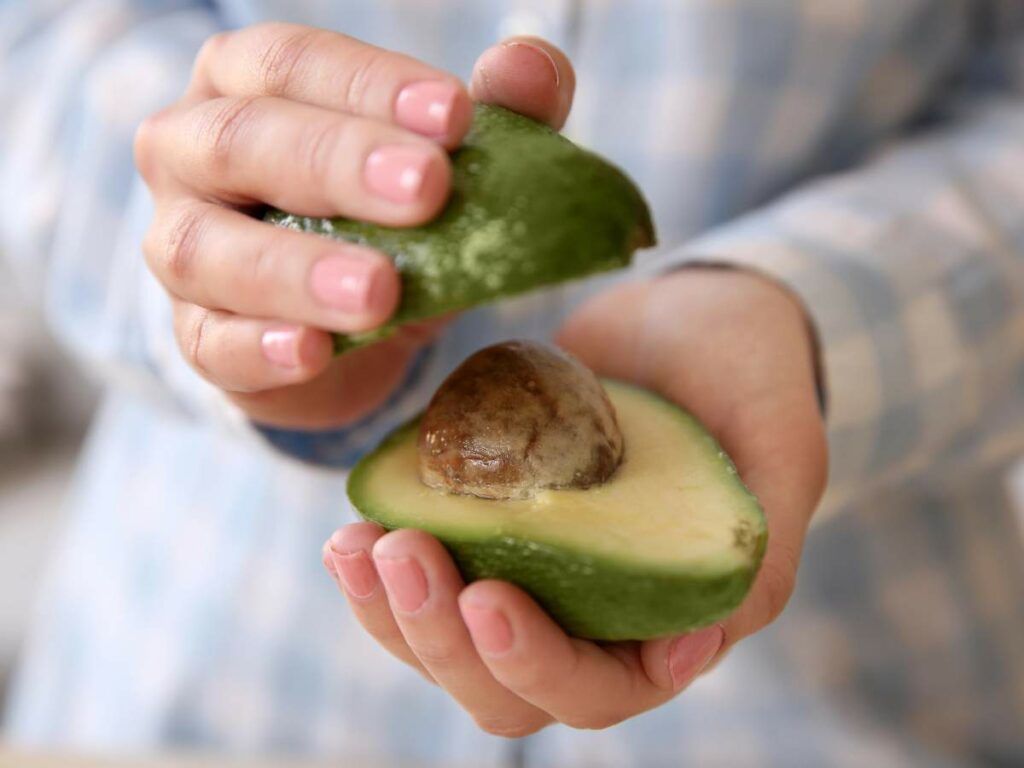 Two hands hold a halved avocado with the seed visible, while wearing a blue checkered shirt.