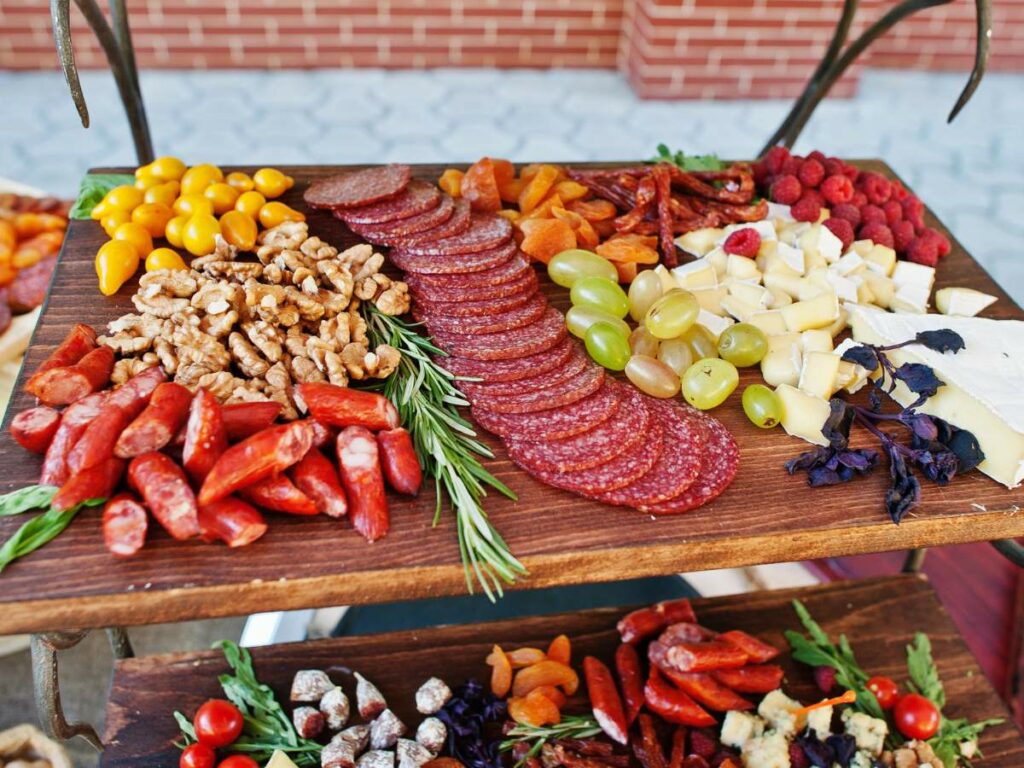 A wooden board topped with salami slices, grapes, nuts, cheese cubes, cherry tomatoes, dried fruits, and sausage chunks.