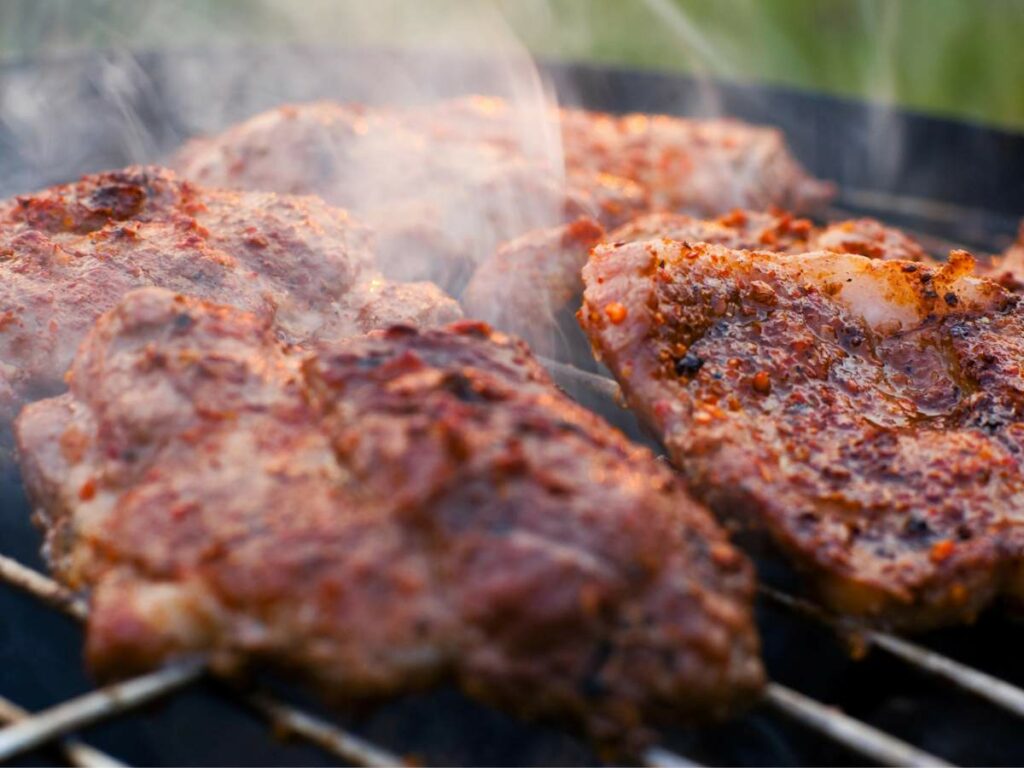 Close-up of seasoned meat pieces being grilled on a barbecue with visible smoke rising.