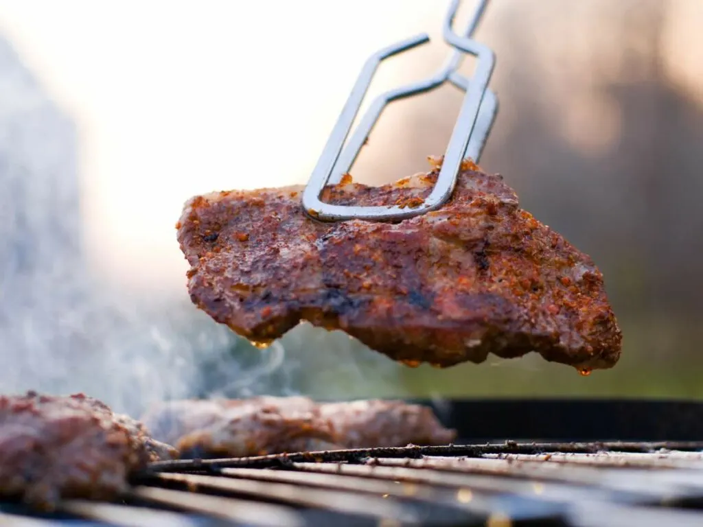 Close-up of a pair of tongs holding a piece of seasoned meat over a grill, with other pieces of meat cooking in the background.