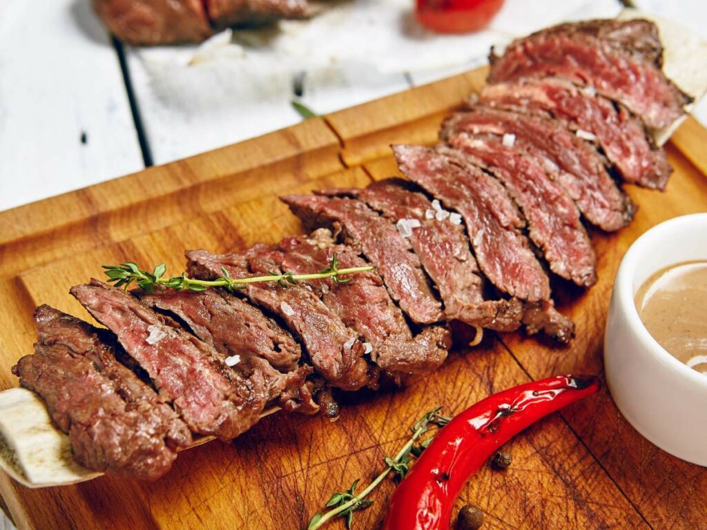 Sliced medium-rare steak on a wooden board, garnished with fresh thyme and a red chili pepper, served with a small bowl of dipping sauce on the side.