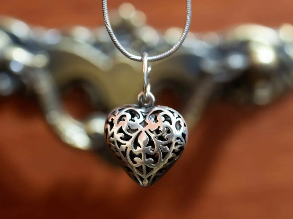 Close-up of a silver filigree heart-shaped pendant on a chain, with a blurred bronze-colored ornate background.