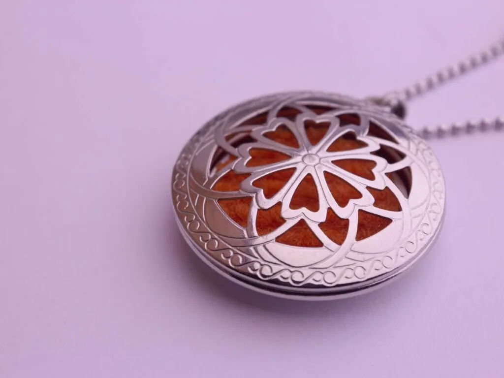 A close-up of a silver pendant with an intricate floral design, featuring an orange background, hanging from a beaded chain on a light purple surface.