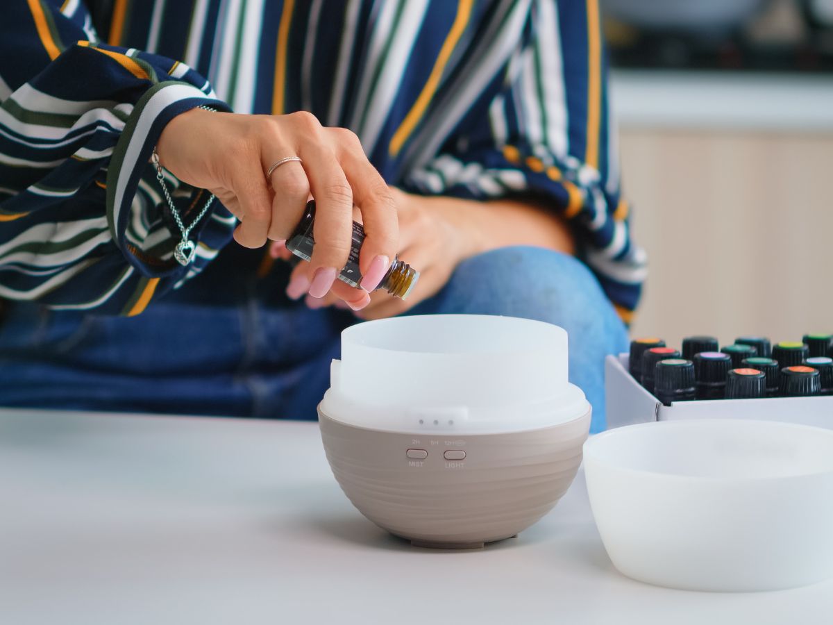 A person wearing a striped shirt is adding essential oil from a small bottle into a diffuser on a white table.