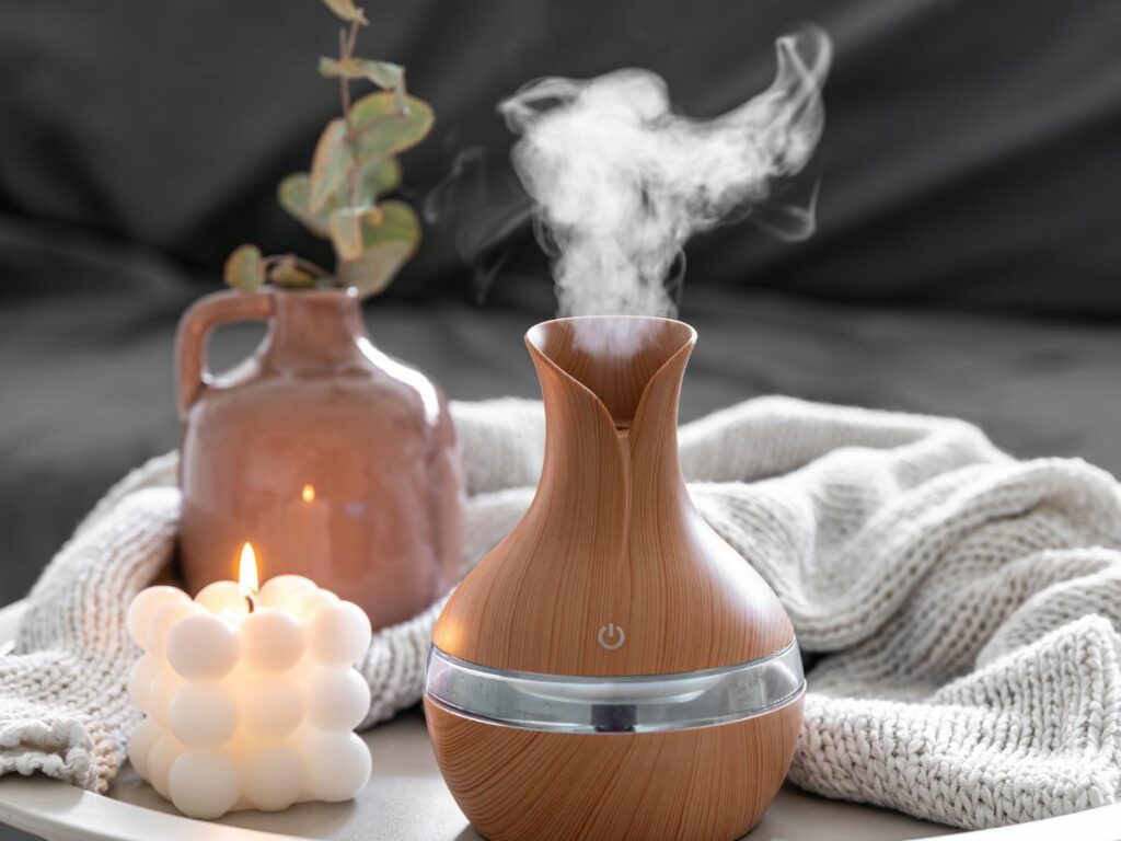 A wooden grain aromatherapy diffuser emitting vapor sits on a table beside a lit bubble candle and a ceramic vase with greenery, with a knitted blanket in the background.