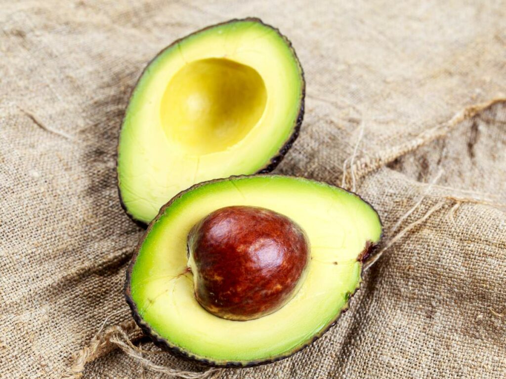 A halved avocado with one side containing the seed and the other side showing a smooth, empty pit rests on a burlap fabric surface.