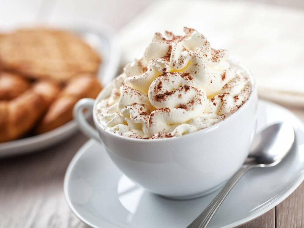 A white cup filled with a whipped cream-topped hot beverage is on a white saucer with a spoon beside it. In the background, some biscuits and croissants are on a plate.