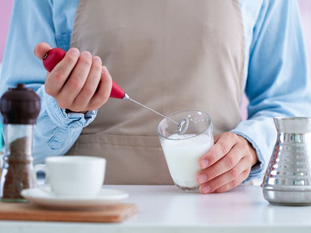 A person wearing an apron uses a handheld milk frother in a glass of milk. A cup, pepper grinder, and metal pitcher are on the table.