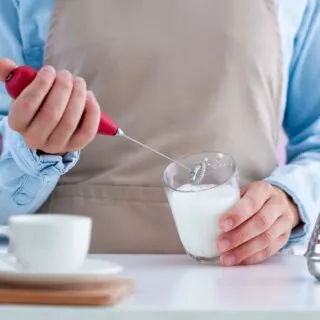 A person wearing an apron uses a handheld milk frother in a glass of milk.