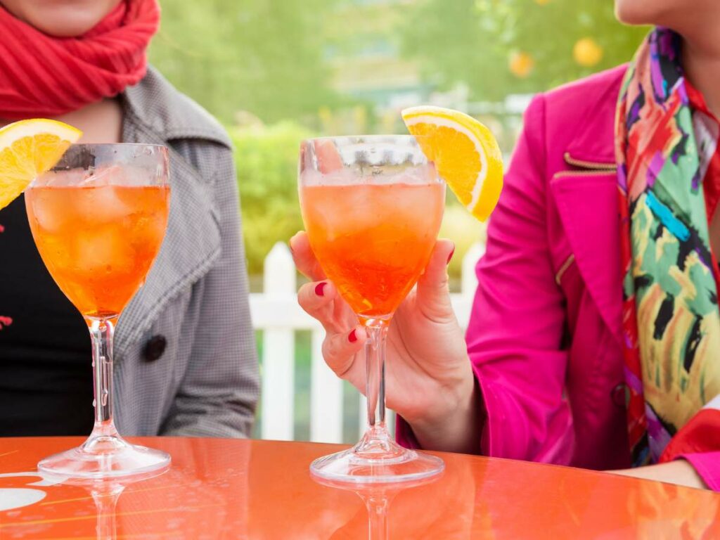 Two people holding wine glasses with orange-colored drinks, garnished with orange slices.