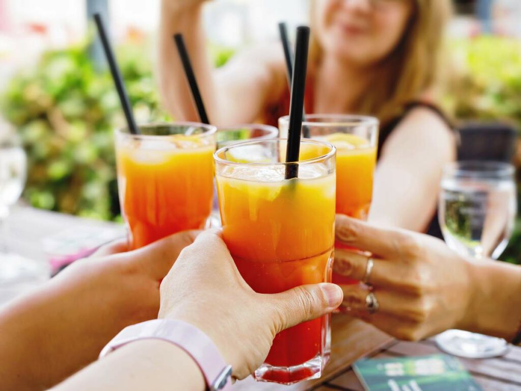 Four people clinking glasses filled with orange and red beverages through black straws in an outdoor setting.