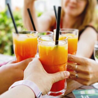 Four people clinking glasses filled with orange and red beverages through black straws in an outdoor setting.