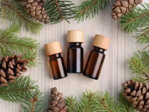 Three small amber bottles with wooden caps are placed in the center of a table, surrounded by pinecones and fir branches.