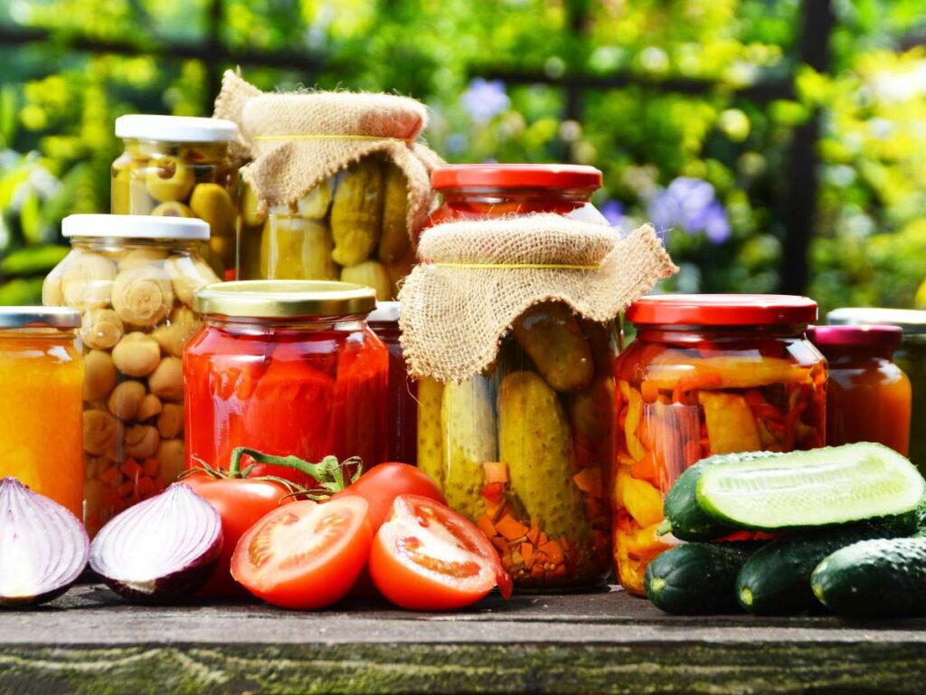 A variety of preserved vegetables in jars, surrounded by fresh produce like tomatoes, cucumbers, and onions, displayed on a wooden surface in an outdoor setting.