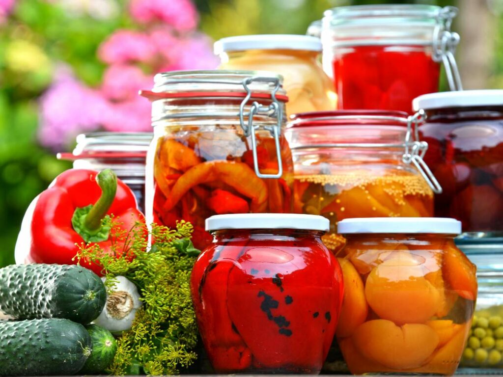 An assortment of pickled vegetables in glass jars, including red peppers and cucumbers, displayed outdoors with fresh vegetables like cucumbers, a red bell pepper, and garlic at the forefront.