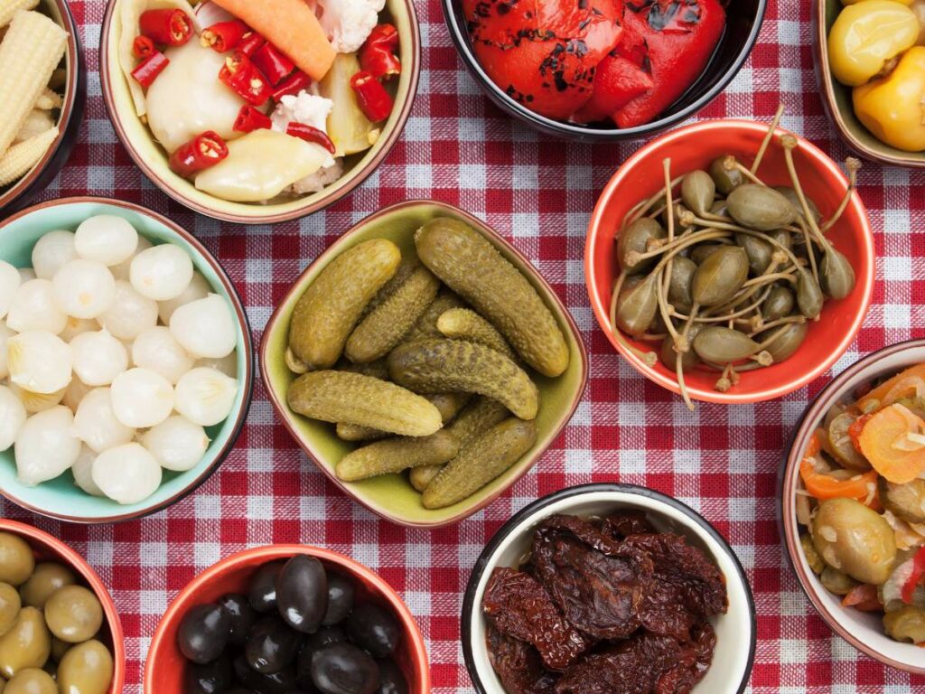 An assortment of pickled vegetables and condiments in small bowls on a red and white checkered tablecloth.