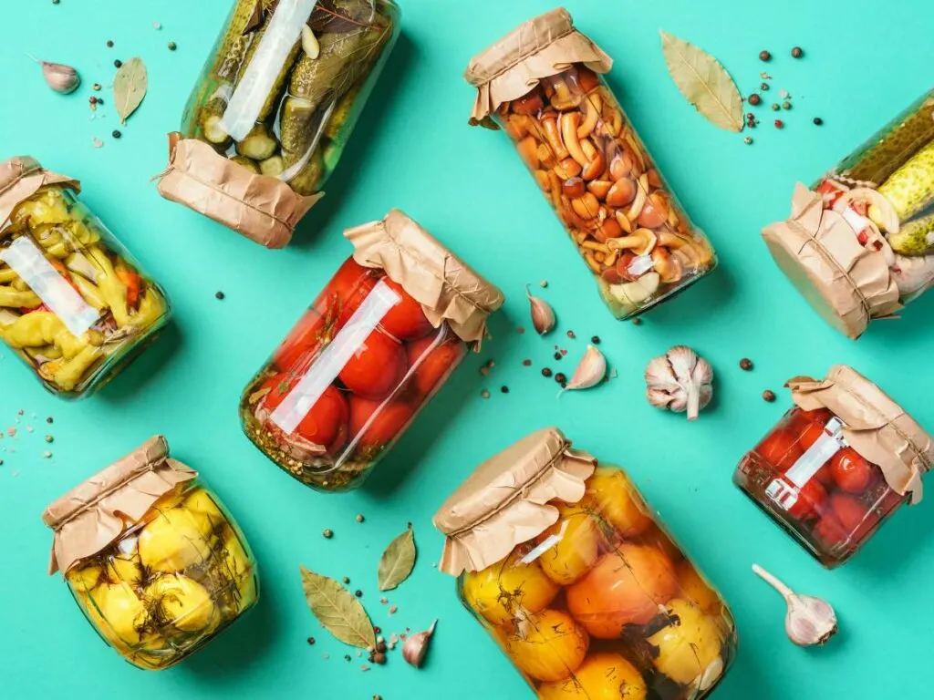Various jars of preserved vegetables, including pickles, tomatoes, and mushrooms, arranged on a turquoise surface with scattered garlic cloves and bay leaves.
