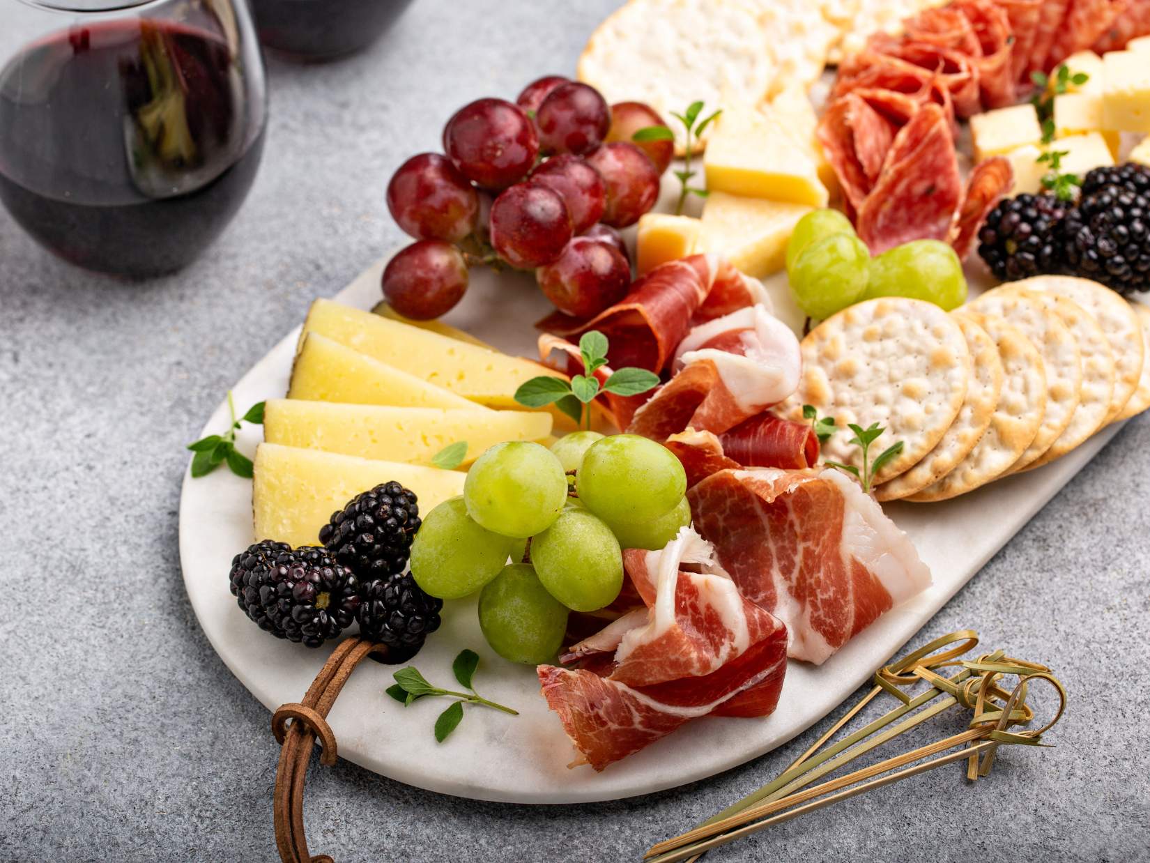 A small charcuterie board with slices of cheese, cured meats, grapes, blackberries, crackers, and herbs.