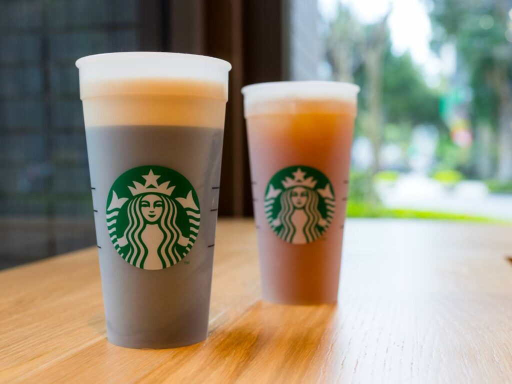 Two Starbucks cups filled with different beverages are placed on a wooden table near a window.