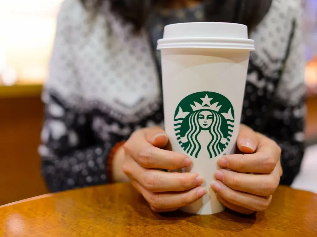 A person holding a white Starbucks cup with a green logo on a wooden table.